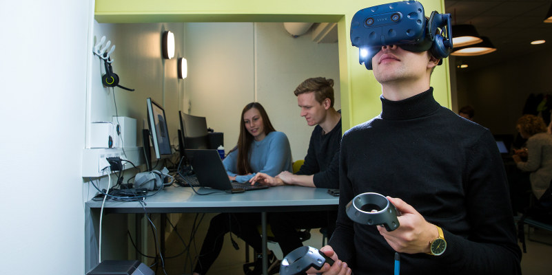 En person med VR-briller og VR-ustyr i hånda og to personer som jobber på en PC i bakgrunnen. Foto.