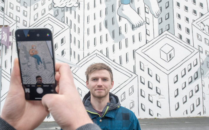 A pair of hands holding a mobile phone which takes a picture of a man who is standing in front of a wall with street art. Photo. 