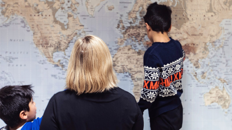 A young child points at a large map, his back towards the camera. A woman and a child sit in the foreground. Photo.