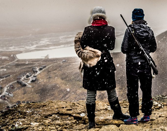 Two people standing on a mountain looking out over a field, one person is carrying a hunting rifle. Photo.