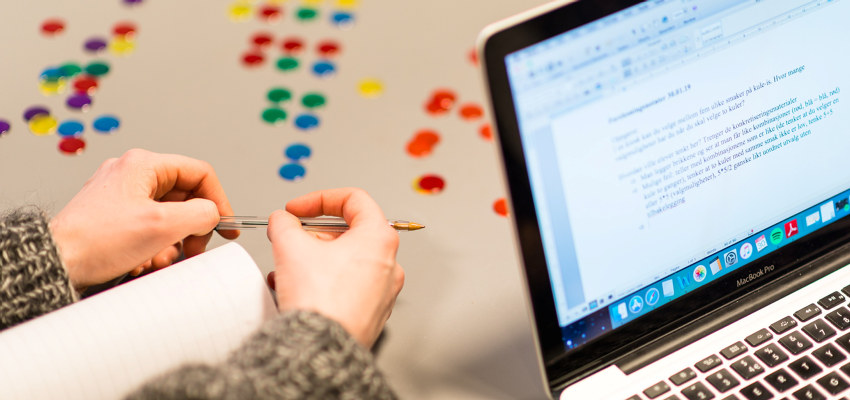 En hånd holder en penn ved siden av en pc og flere knapper i ulike farger. Foto.