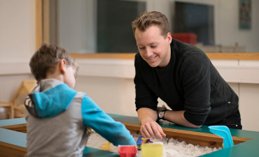 Et barn leiker i ei sandkasse saman med en voksen. Foto.