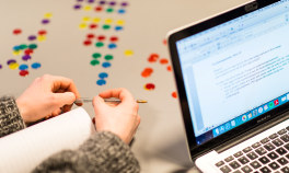 Hender som holder en penn. En laptop med tekst. Runde brikker i ule farger som ligger bordet i mønster. Foto