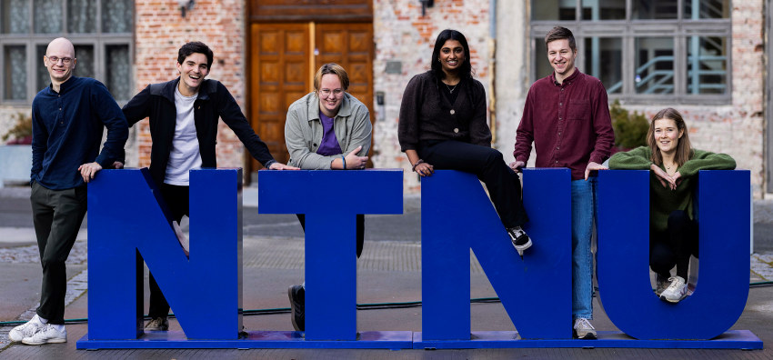 Seks studenter står ved store fysiske NTNU-bokstaver utendørs. Foto