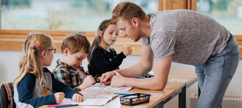 En lærerstudent står i et klasserom ved pultene til tre elever, to jenter og en gutt. De jobber med oppgaver i bøker. Lærerstudenten peker i boken til eleven og forklarer. Foto