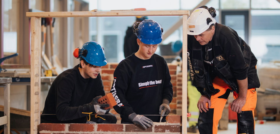 To elever og en lærer på et verksted, bygger en mur. Alle har på arbeidsklær. Foto