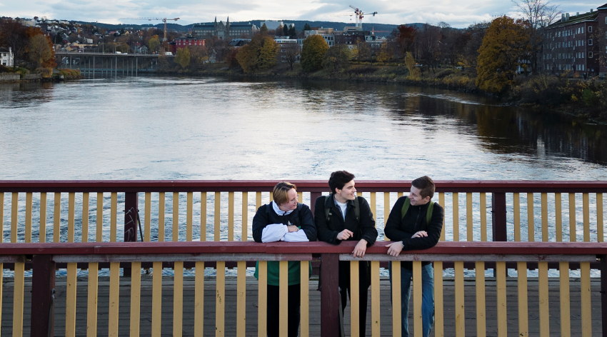 Tre studenter står på en bru over Nidelven, en dame og to menn. Foto