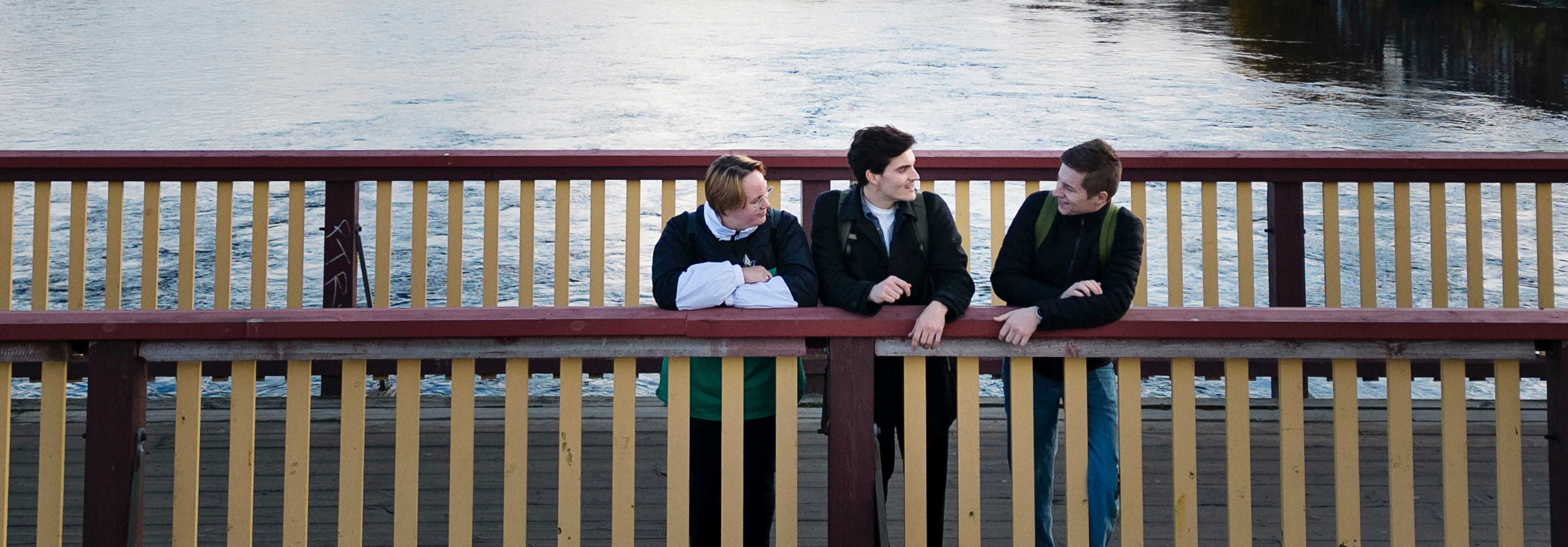 Three people stand on a wooden bridge with red and yellow railings, looking out over calm water. They are leaning on the railing and appear to be talking. The water is visible beneath the bridge. Photo