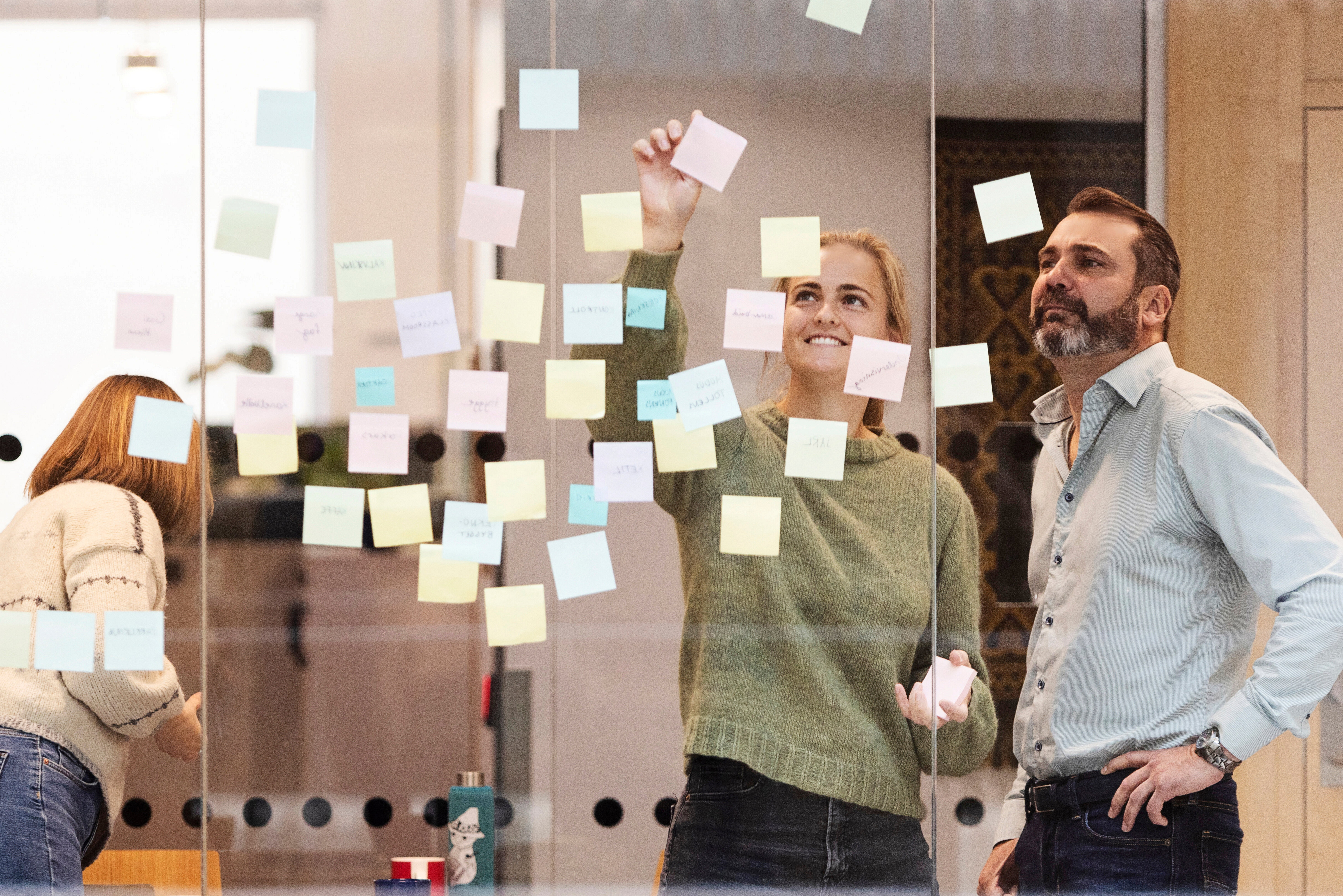 Tre personer står foran en glassvegg dekket med fargerike post-it-lapper. To personer er i fokus, og én av dem rekker opp hånden for å feste en ny lapp. Den tredje personen er delvis synlig til venstre i bildet, vendt bort fra kameraet. Foto