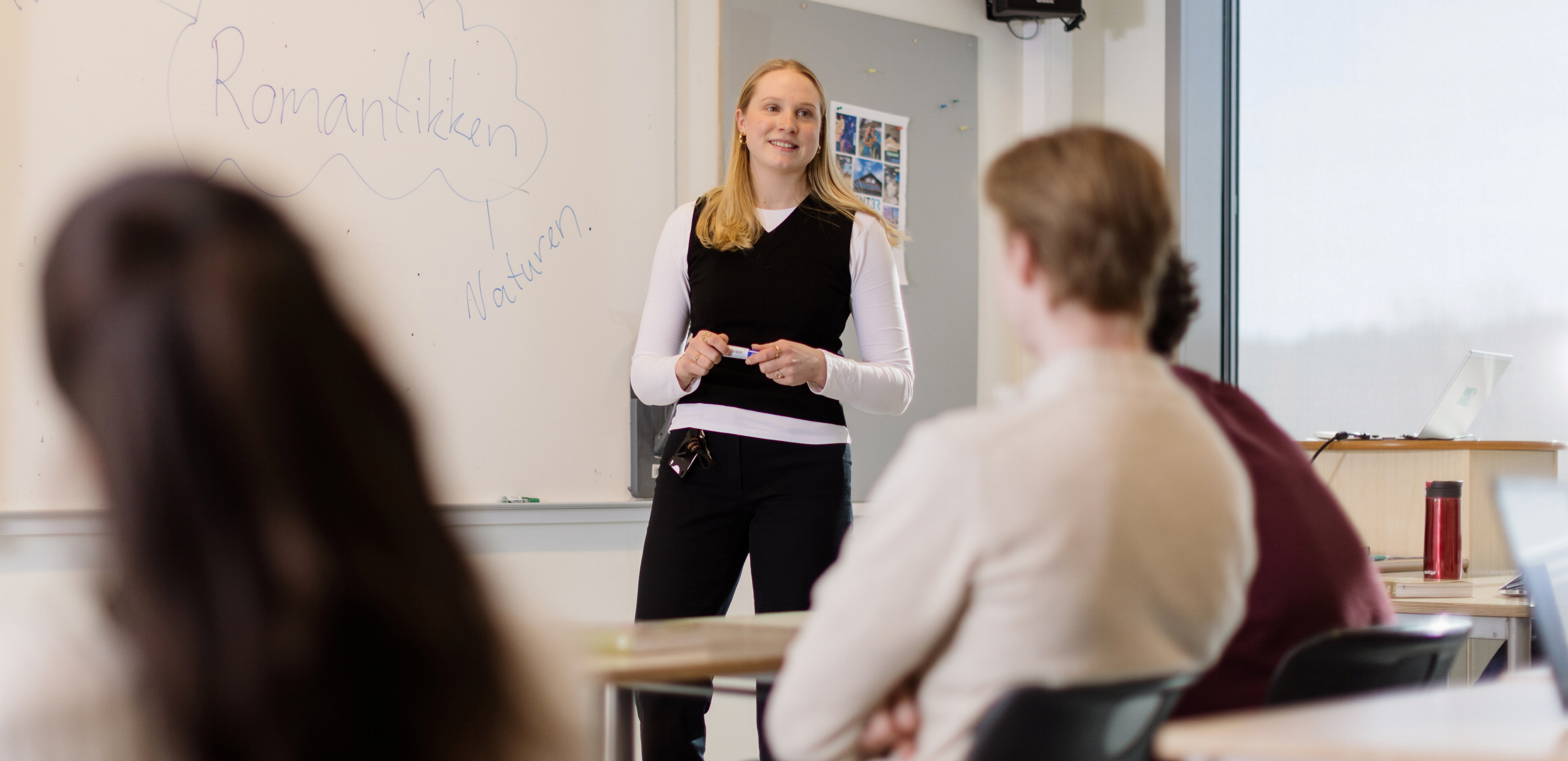 En lærerstudent står foran en tavle og underviser en gruppe elever i et klasserom. På tavlen står det skrevet 'Romantikken' med en pil som peker mot ordet 'Natur'. Elevene sitter ved pulter og følger med på undervisningen. Foto