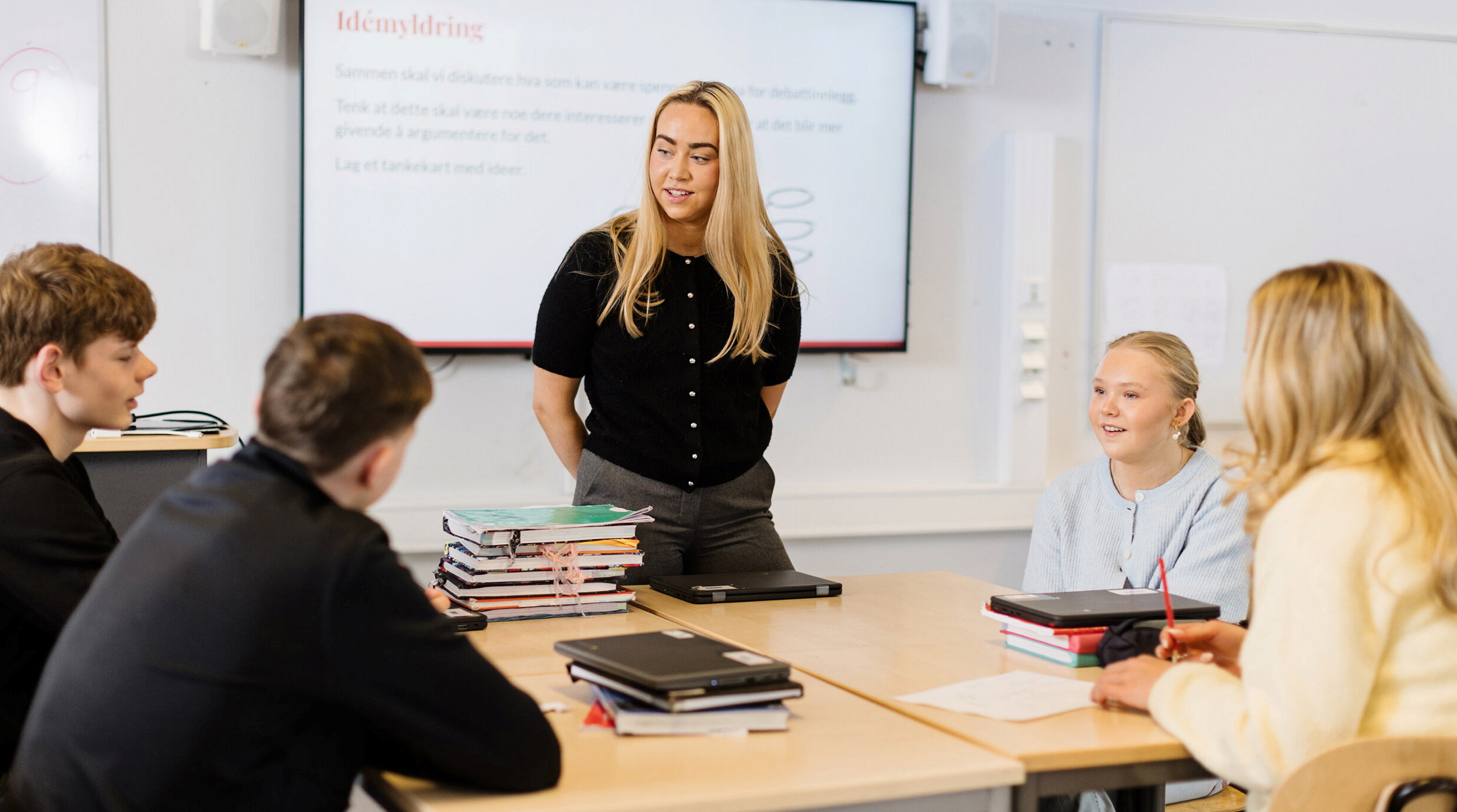 En gruppe elever deltar i en undervisningssituasjon. En lærerstudent står foran en skjerm med teksten "Idemyldring" og gir instruksjoner til de andre, som sitter rundt et bord med bøker og notatbøker. Skjermen viser en oppgave som oppfordrer deltakerne til å lage et tankekart med ideer basert på egne interesser og engasjement. Foto
