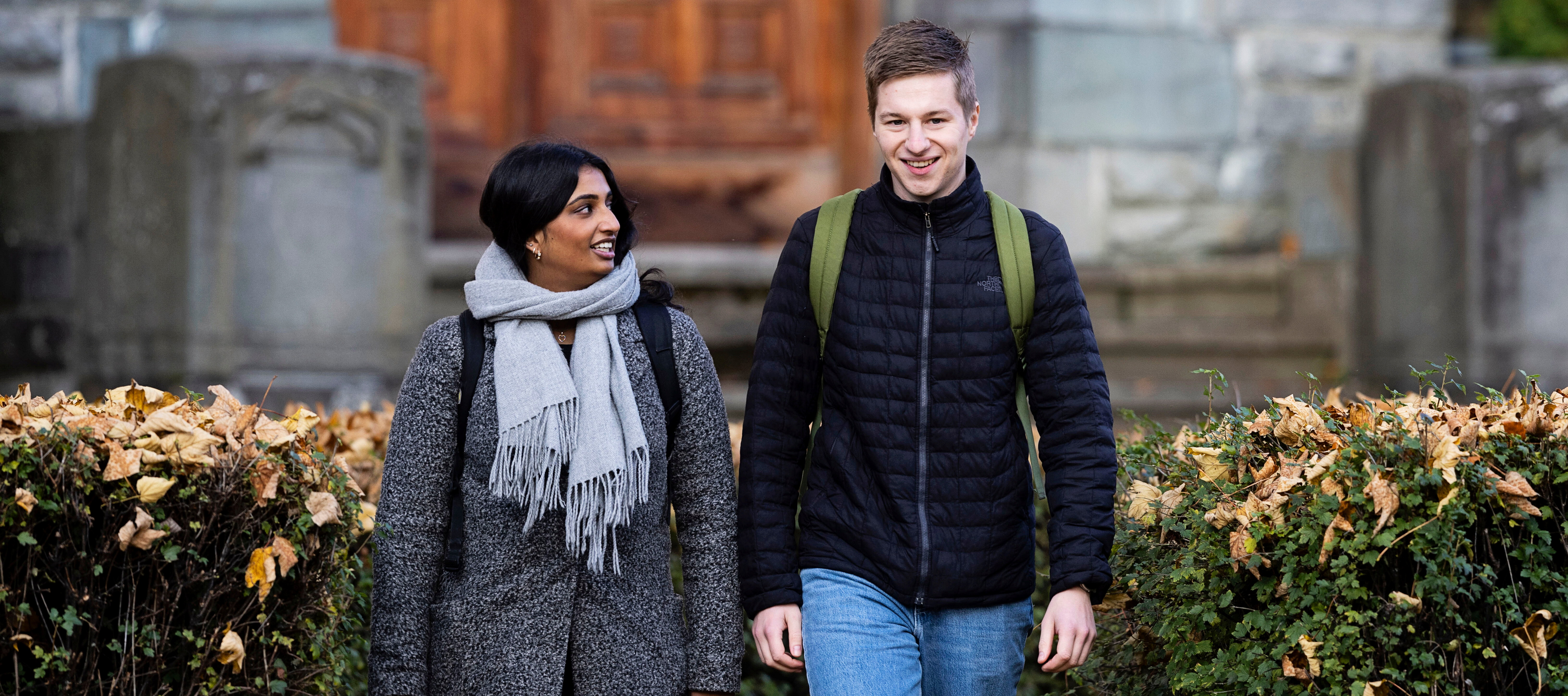 To personer går sammen utendørs foran en bygning med steintrapper og en stor tredør. Den ene personen har på seg en grå frakk og et grått skjerf, mens den andre har en svart jakke og blå jeans. Begge bærer ryggsekker. Foto