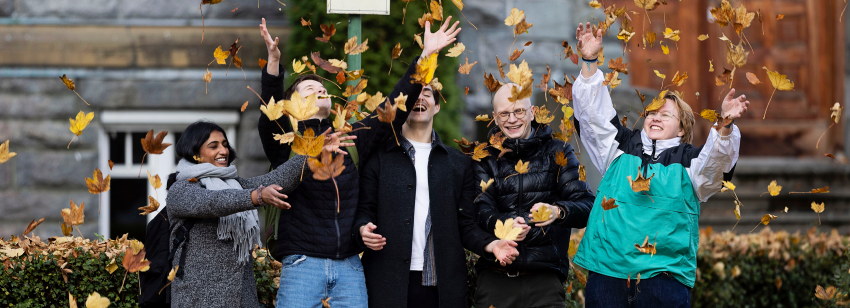 Fem personer står foran en bygning med steinfasade og stor treport, og kaster gult og oransje høstløv opp i luften. De smiler og ser ut til å ha det gøy sammen. Foto
