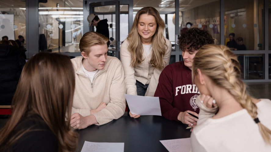 A group of teacher‑education students sit around a table discussing a sheet of text in a bright, modern learning environment. Photo.