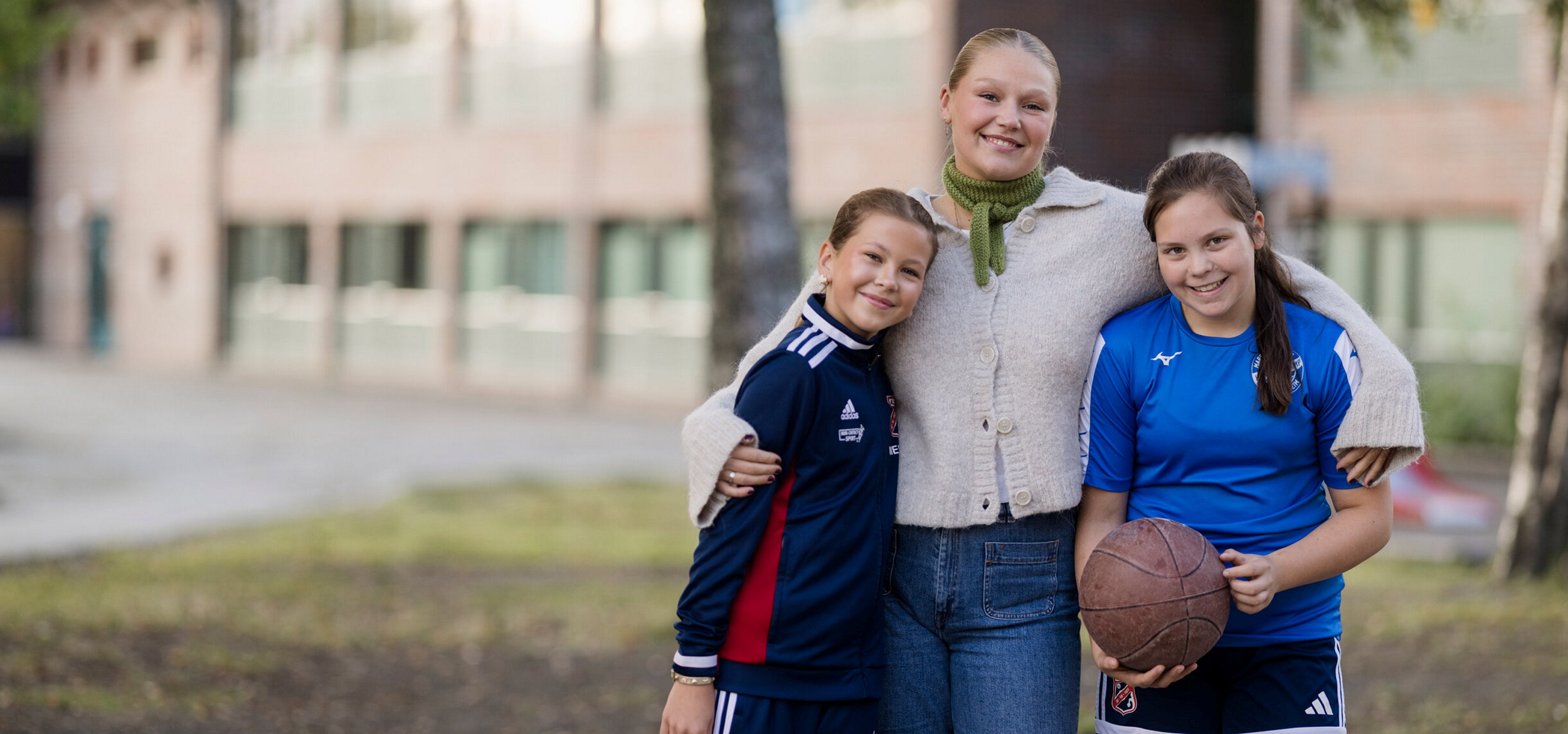 An adult and two children stand together outdoors in front of a school building. The child on the right is holding a basketball, and the three are standing close as a group. Photo.