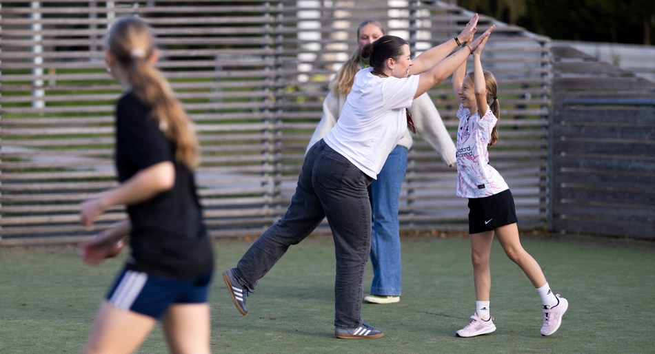 En lærerstudent og en elev står på en utebane og gir hverandre en «high five», mens andre barn løper og spiller i bakgrunnen. Scenen viser aktivitet og lek i et utemiljø. Foto