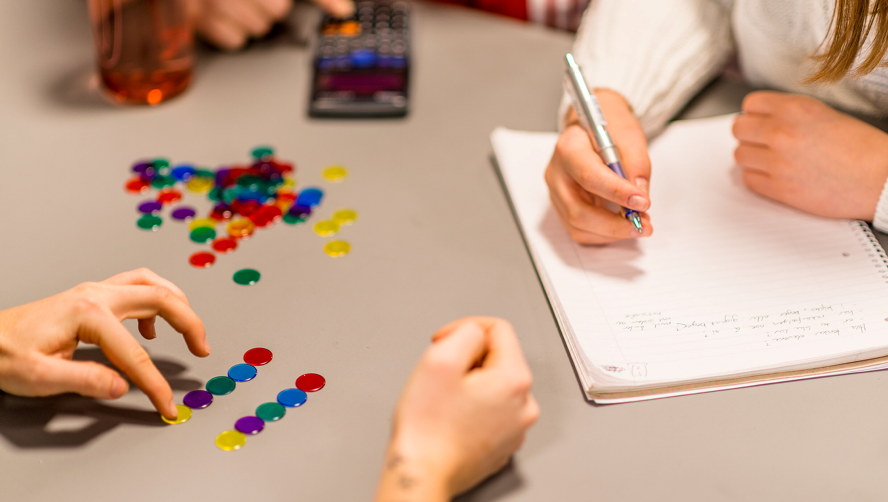 Hands working at a table with colored pieces. One person is writing in a notebook while others sort or move the pieces. Photo.