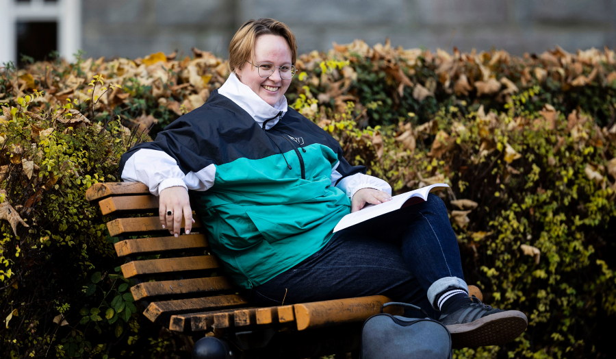 En person sitter avslappet på en trebenk utendørs, omgitt av busker med høstfargede blader. Personen har på seg en grønn og svart jakke og mørke jeans, og holder en åpen bok i fanget. Foto