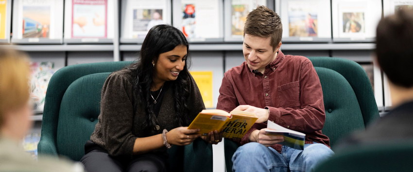 To lærerstudenter sitter i dype, grønne stoler i et bibliotek eller leseområde og ser i en bok sammen. Hyller med bøker og tidsskrifter er synlige i bakgrunnen, og to andre lærerstudenter sitter i forgrunnen med ryggen til kameraet. Foto