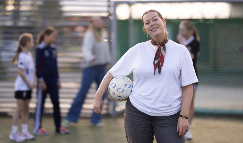 Person står på en idrettsbane og holder en fotball under armen. I bakgrunnen er flere personer og et målområde synlig, delvis ute av fokus. Foto